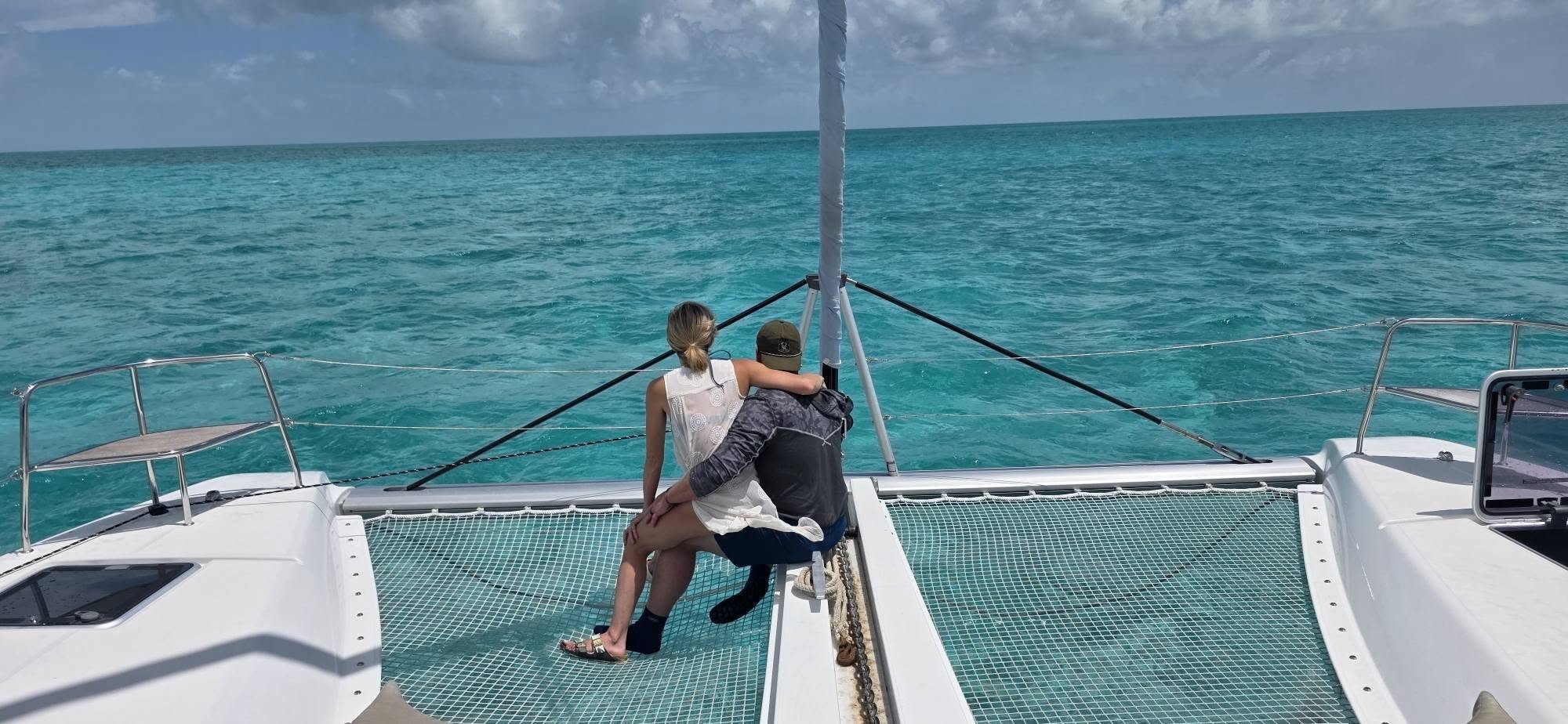 A couple sitting together on a catamaran bow overlooking turquoise water